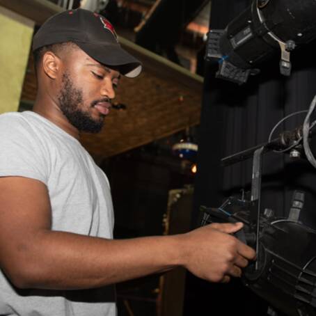 Person adjusting stage lighting backstage, with another person partially visible in the background.