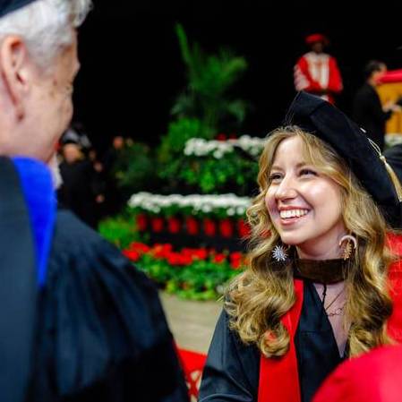 A smiling graduate in a black cap and gown with a red stole interacts at a graduation ceremony.
