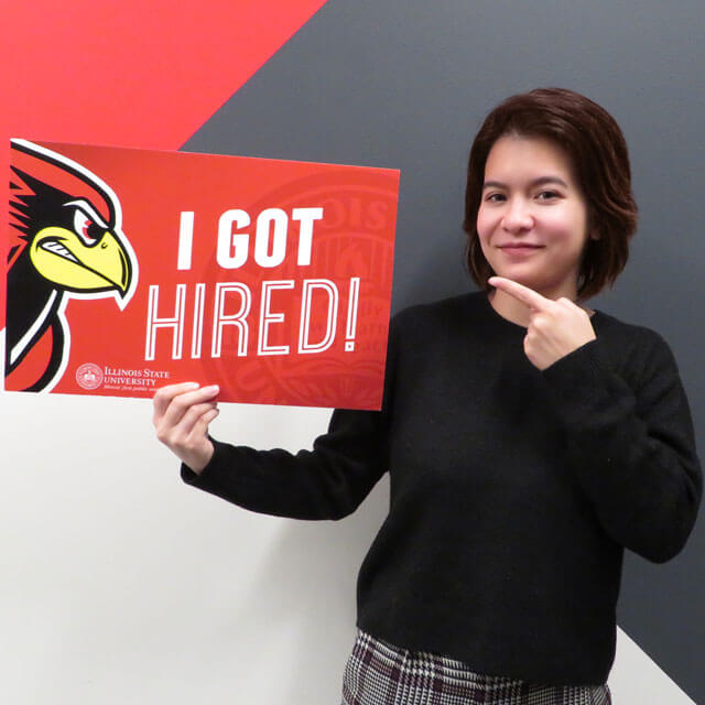 A person holds and points to a sign that reads 'I got hired' with a Reggie Redbird logo.